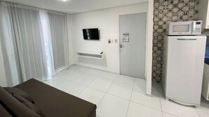 A room with a flat-screen TV mounted on the wall, a white refrigerator with a microwave on top, a door with a sign, and a window with curtains. - Porto Sol Flats by AFT (Ipojuca)