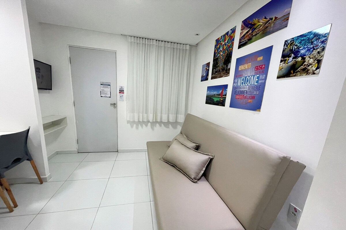A room with a beige sofa, a small desk, and a television on the wall. There are several posters on the wall, and a door with a sign on it.