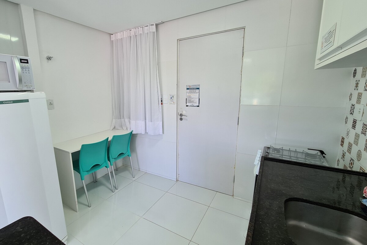 A small, clean kitchenette with a microwave, a refrigerator, a sink, and a window with curtains.
