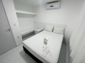 Una habitación de hotel limpia y blanca, con una cama, una mesita de noche pequeña, aire acondicionado de pared y una puerta.