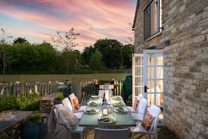 Un área de comedor al aire libre con una mesa dispuesta para seis personas, rodeada de muros de piedra y una valla de madera.