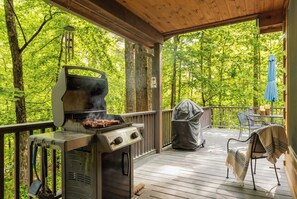 An outdoor grill area with a barbecue, a covered grill, and a table with chairs on a wooden deck surrounded by trees. - Camp Sasquatch*3B/3BA*PetsOk*Pool (Deep Gap)