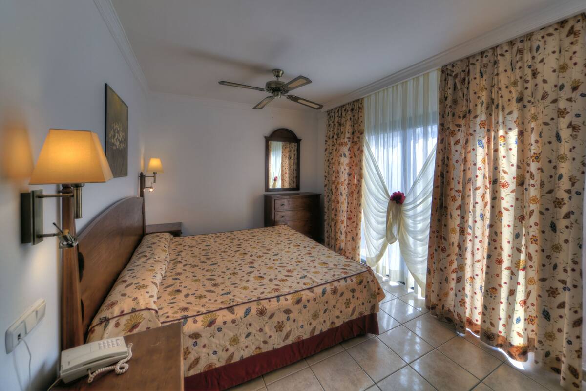 A hotel room with a bed, a ceiling fan, a nightstand with a lamp, a telephone, and a window with curtains.