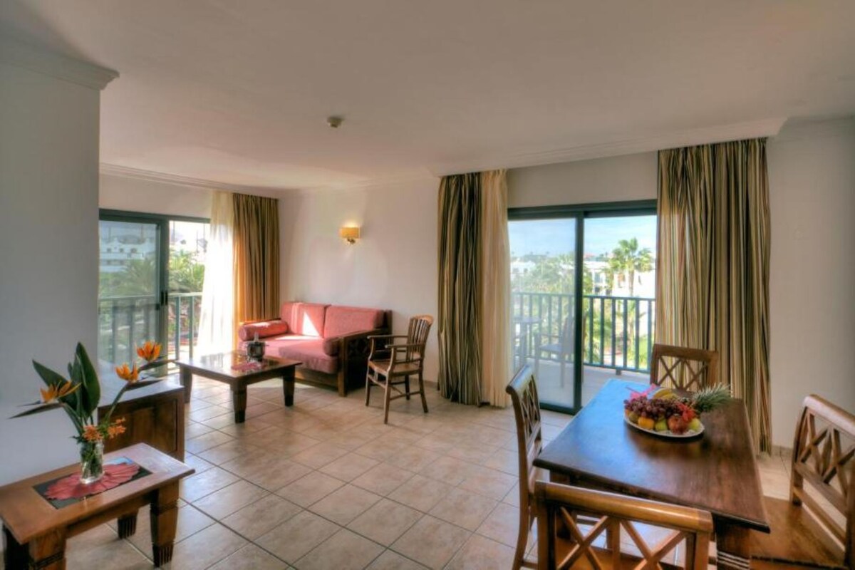 A hotel room with a balcony, a sofa, a dining table with chairs, and a small table with a vase of flowers.