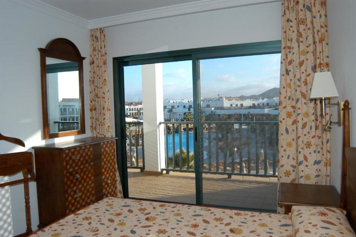 A hotel room with a balcony, a bed, a dresser, a mirror, and a view of the city and sea.