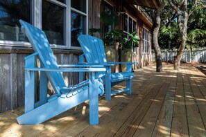 Zwei blaue Adirondack-Stühle auf einer Holzterrasse mit Blick auf ein Haus und Bäume.