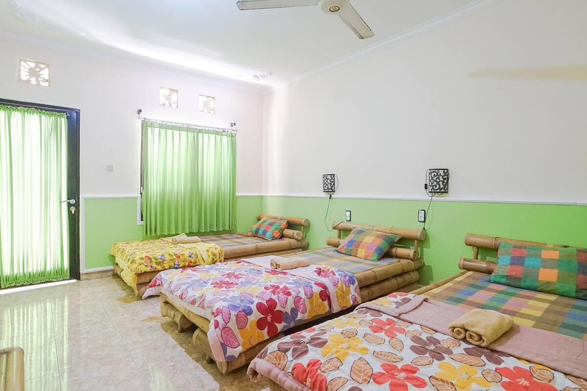 Quarto com três camas, cada uma com colcha florida, cortinas verdes e ventilador de teto.