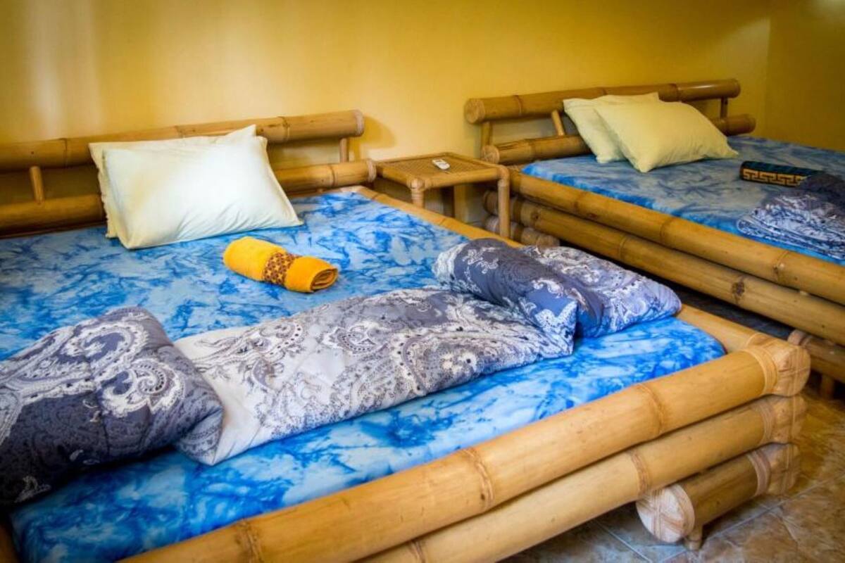 Quarto com duas camas, cada uma com colcha e almofadas estampadas em azul, uma pequena mesa de cabeceira de madeira e uma toalha amarela.