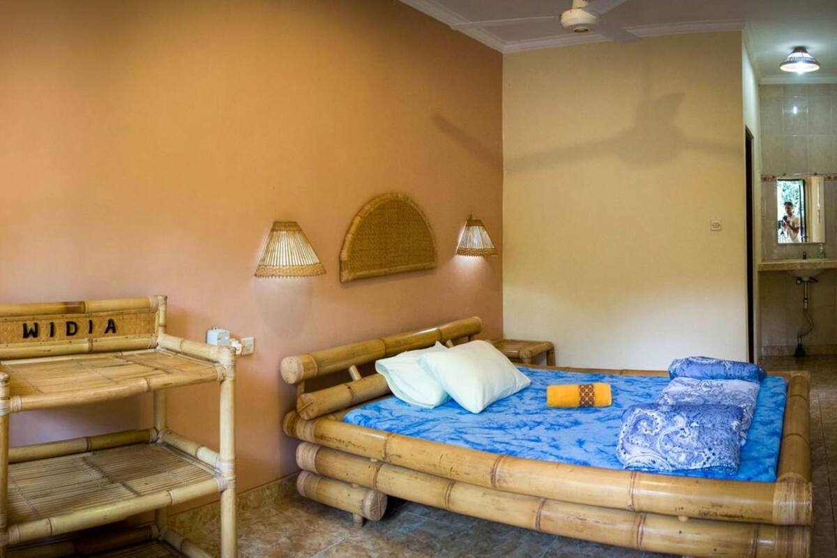 Quarto com cama de bambu, um banco com a inscrição 'WIDIA' e duas luminárias fixadas na parede.