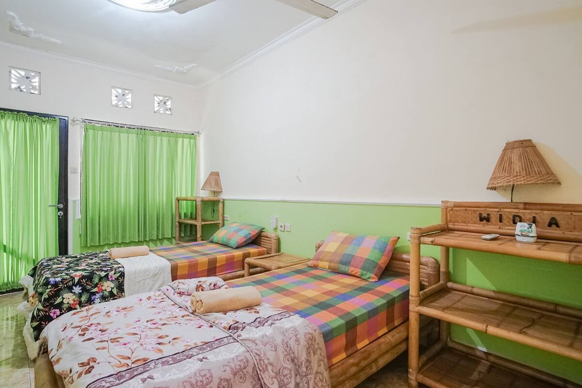 Quarto com duas camas, uma cortina verde, uma prateleira de madeira com abajur e um letreiro fixado na parede.