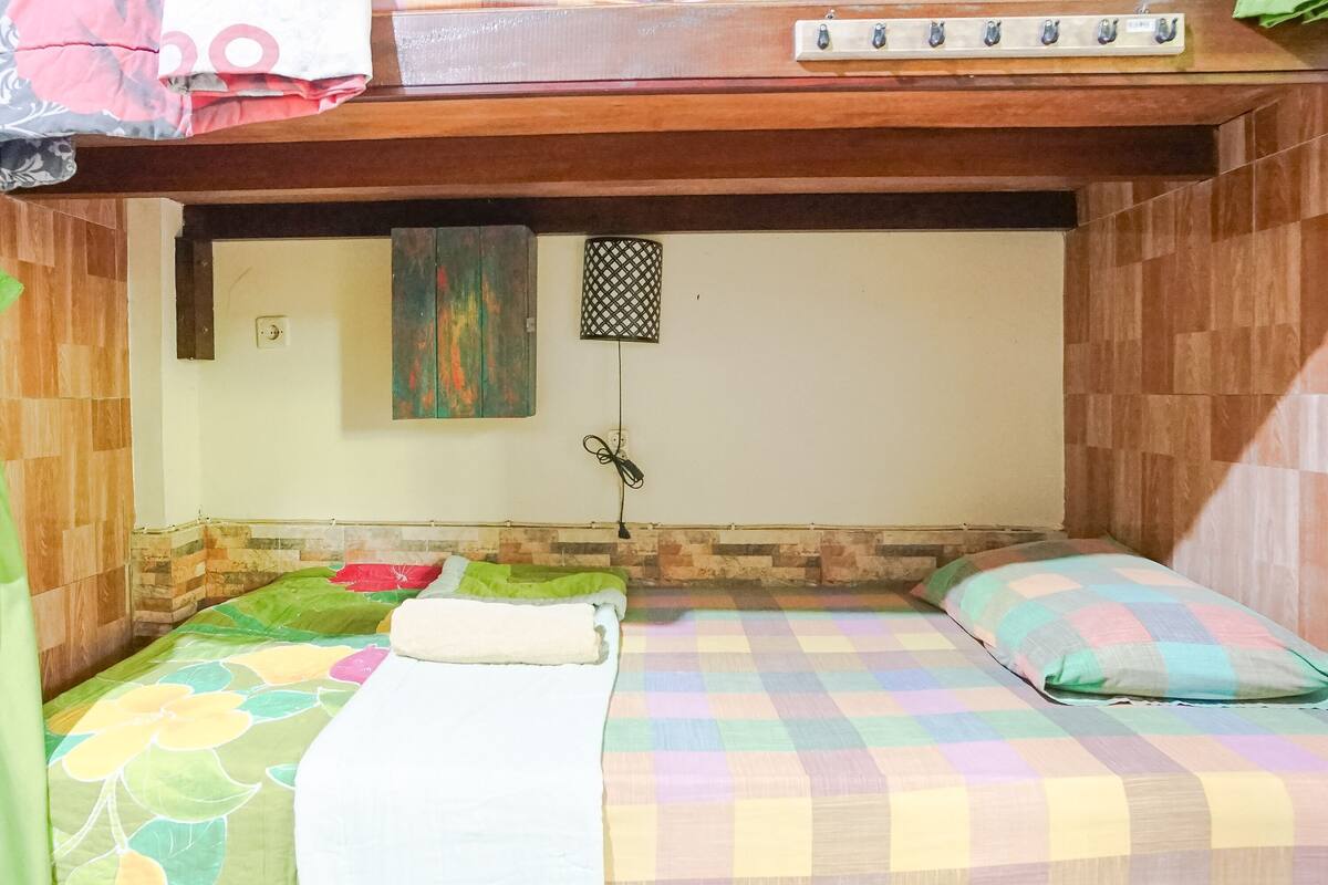 Quarto pequeno e acolhedor com duas camas, cada uma com colcha colorida. Há uma cabeceira de madeira acima das camas, uma pequena prateleira com ganchos e um aplique de parede.