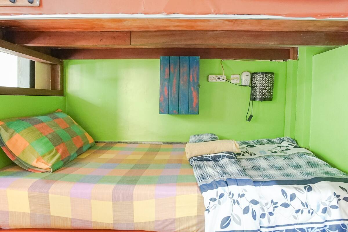 Um quarto pequeno com paredes verdes, uma cama de solteiro, uma janela e uma tomada na parede.