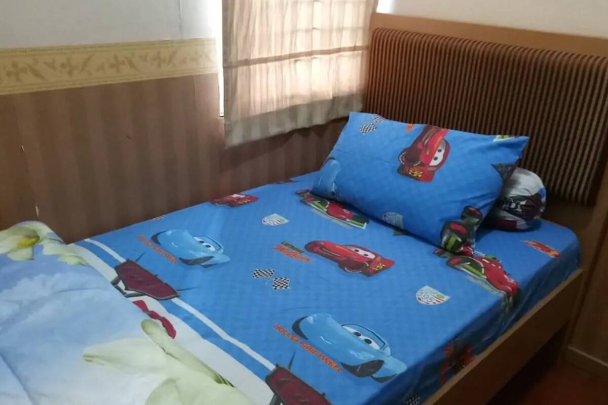 Un lit simple avec une couvre-lit bleue ornée de motifs de voitures de dessins animés et un oreiller présentant un motif similaire.