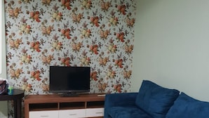 Un salon avec un papier peint à motifs floraux, un canapé bleu, un meuble de rangement en bois avec un téléviseur et un sol carrelé.