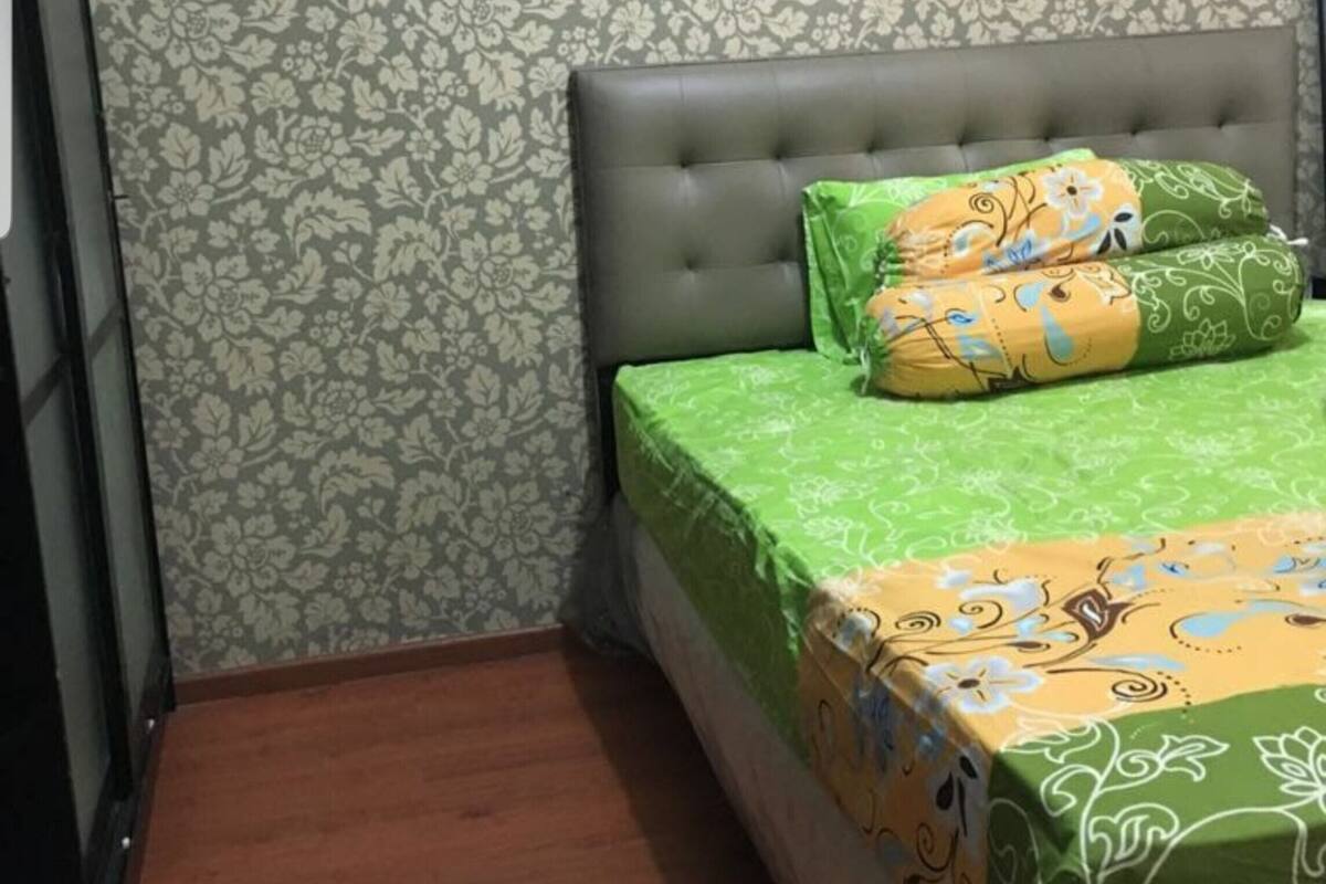 Une chambre à coucher avec une tête de lit capitonnée grise, un papier peint à motifs floraux et une couvre-lit verte ornée de motifs floraux.
