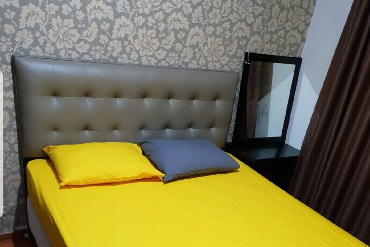 Un lit avec une tête de lit rembourrée de couleur grise, un protège-matelas jaune et deux oreillers, un jaune et un bleu.