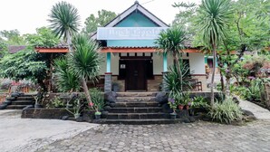 A building with a sign reading "Rock Garden." It has a tiled roof, a front entrance with steps, and is surrounded by various plants and trees.