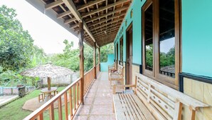 A covered outdoor area with wooden benches and a thatched umbrella, surrounded by greenery.