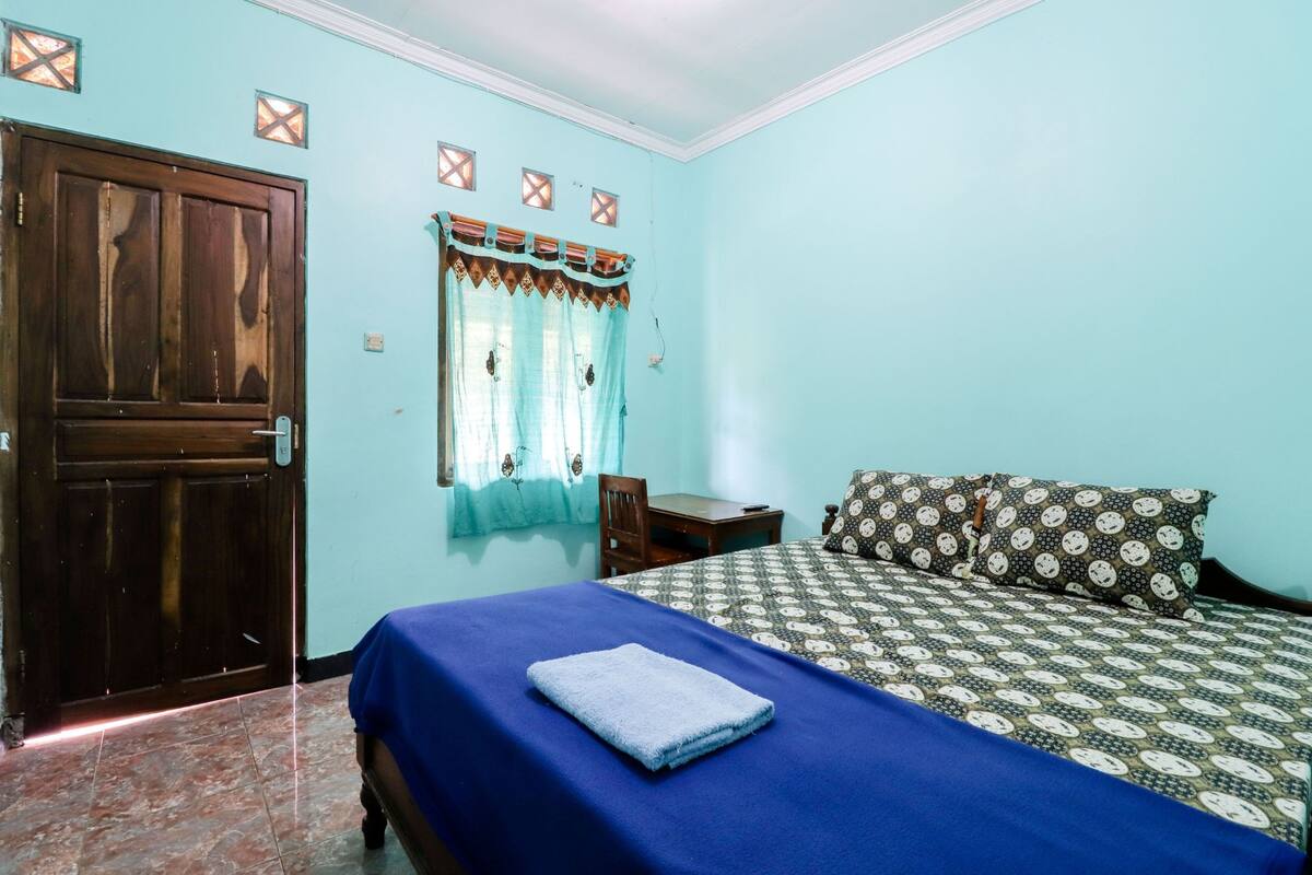A bedroom with a bed, a wooden door, a small table, and a window with curtains.