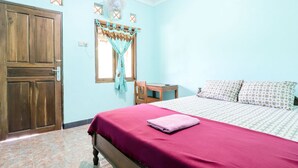 A bedroom with a wooden door, a bed with a red bedspread, a wooden table, and a window with curtains.