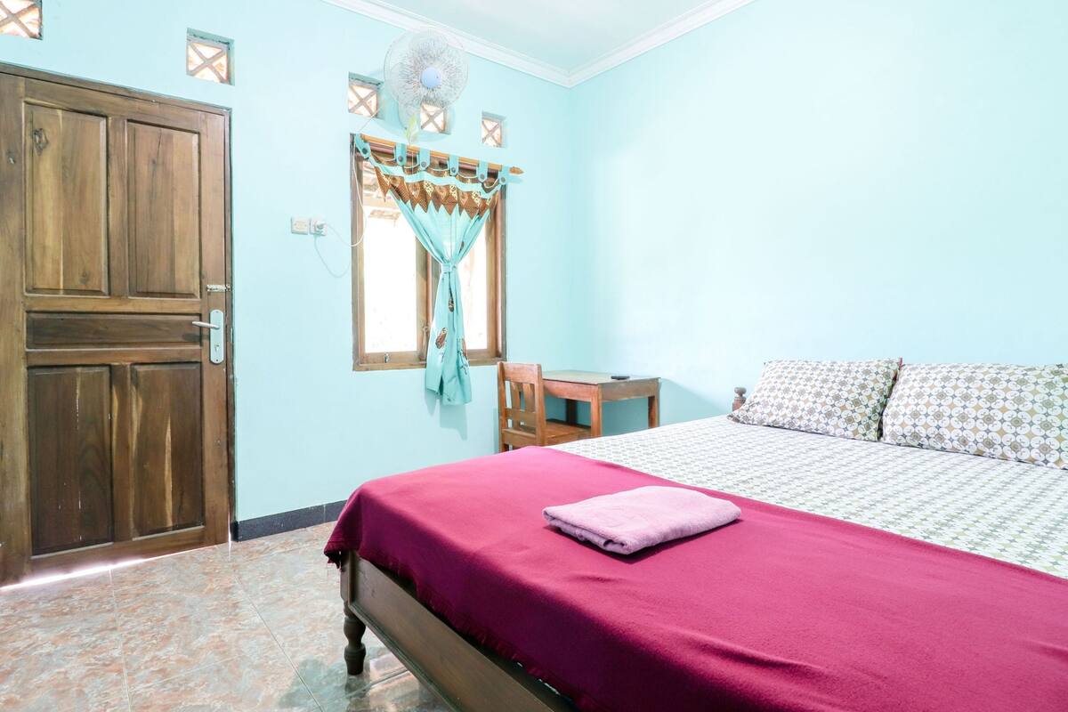 A bedroom with a wooden door, a bed with a red bedspread, a wooden table, and a window with curtains.