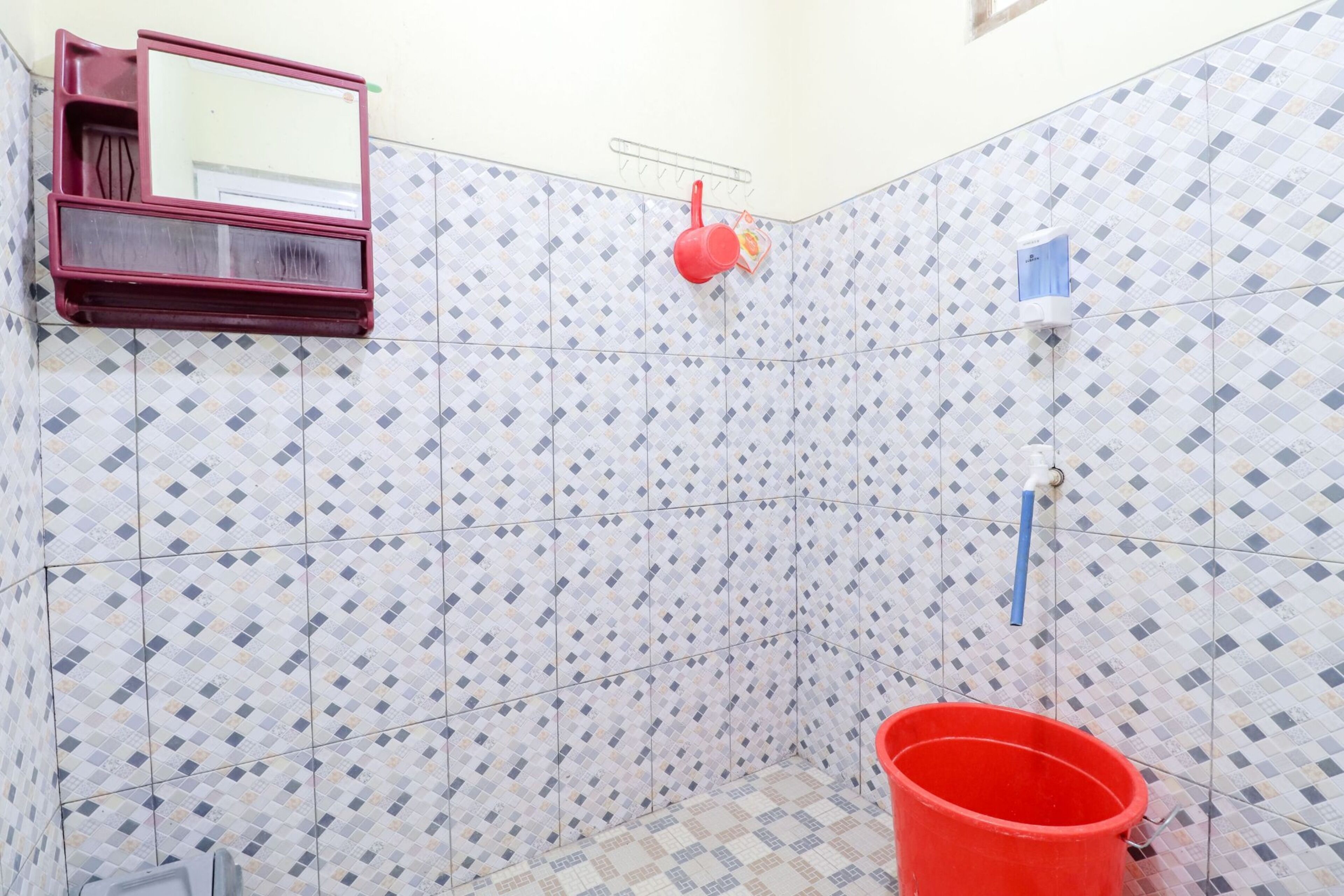 A bathroom with patterned tiles, a red bucket, a mirror, and a soap dispenser.