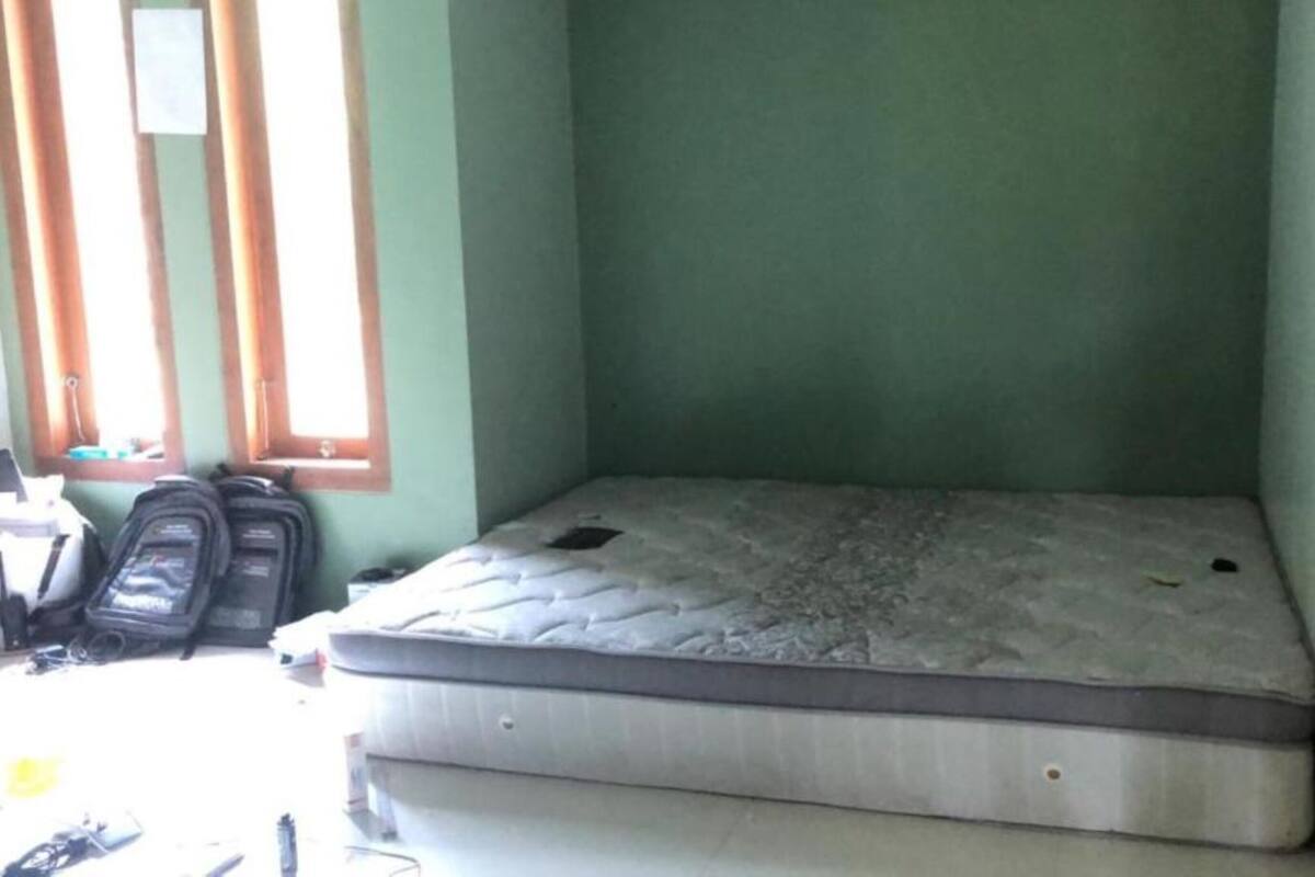 A small, single-bed room with a mattress, a wooden door, and a window with blinds.
