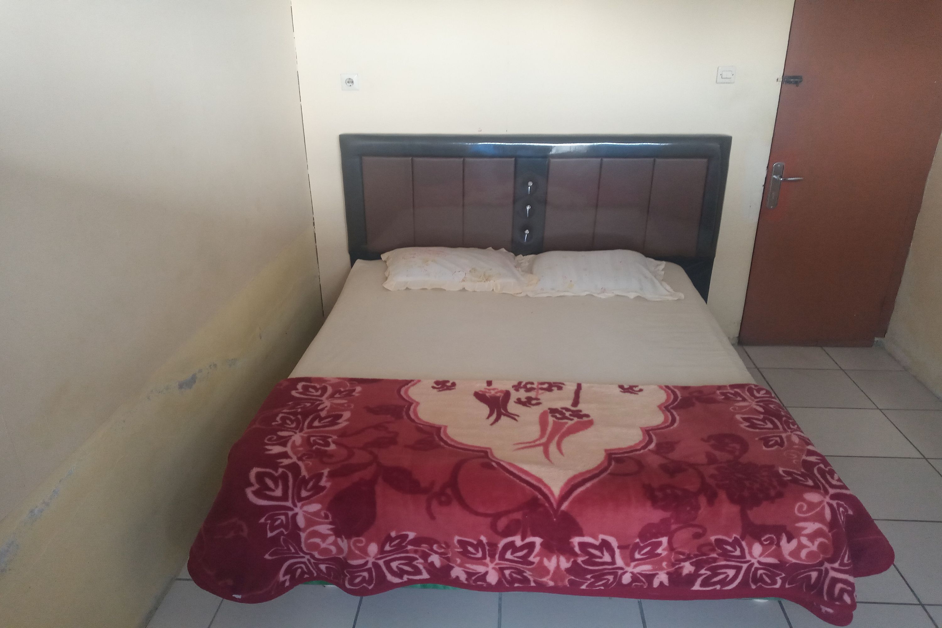 Una cama pequeña y bien hecha con una colcha estampada y almohadas, un cabecero de madera y una puerta cerrada a la derecha.