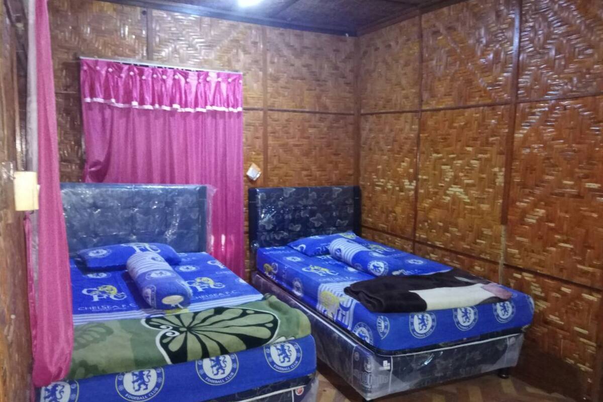 Ein Zimmer mit zwei Betten, beide mit Bettzeug des FC Chelsea bezogen. Das Zimmer hat Holzwände und einen rosafarbenen Vorhang.