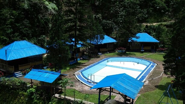 Zona de piscina con una piscina rectangular, rodeada de edificios con techos azules, en un entorno verde y exuberante.