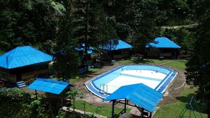 Zona de piscina con una piscina rectangular, rodeada de edificios con techos azules, en un entorno verde y exuberante.