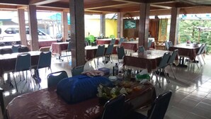 Un restaurante con varias mesas y sillas, cubierto por un techo y con grandes pilares que sostienen la estructura.