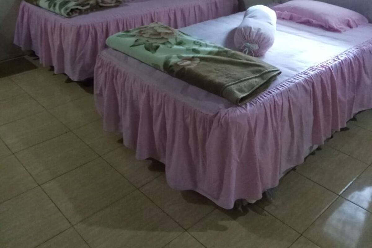 Ein Zimmer mit zwei Einzelbetten, jeweils mit einer rosa Bettdecke und einer blumigen Decke. An der Wand hängt ein gerahmtes Bild und es gibt ein Fenster mit Vorhängen.