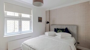 A bedroom with a large window, a radiator, a bed with white bedding, a headboard, and a wall-mounted lamp.
