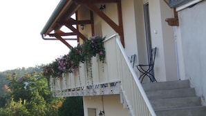 Een balkon met houten hekwerk en bloembakken, een trap en uitzicht op een bebost gebied.
