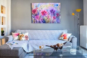 A modern living room with a grey sofa, floral cushions, a glass coffee table, and a colorful abstract painting on the wall.