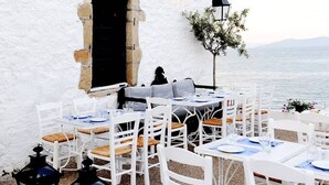 An outdoor dining area with white tables and chairs, a small tree, and a view of the sea. - Thalassa, Return To Timeless Spetses (Spetes)