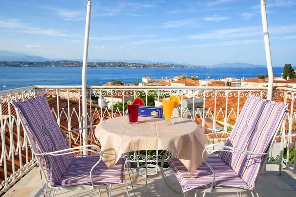 A terrace with two striped chairs, a table with a drink, and a view of the sea and buildings.