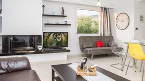 A modern living room with a flat-screen TV, grey sofa, yellow chair, and a dining table with a clock on the wall. - Nina's Villa, Cosy Seaview Retreat (Paralia Vergas)