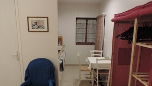 A room with a blue chair, a dining table with chairs, a red coat rack, and a window with patterned curtains. - Independent studio 25m² 800 m from the sea (Anneville-sur-Mer)