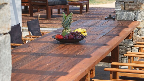A wooden dining table with a fruit bowl, surrounded by wooden chairs, under a stone wall and a pergola with hanging lights. - Charming little villa Elena with private pool and sea views! (Paros)