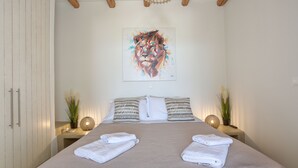 A neatly made bed with two towels, a geometric painting on the wall, and a bedside table with a lamp and a plant. - Charming little villa Elena with private pool and sea views! (Paros)
