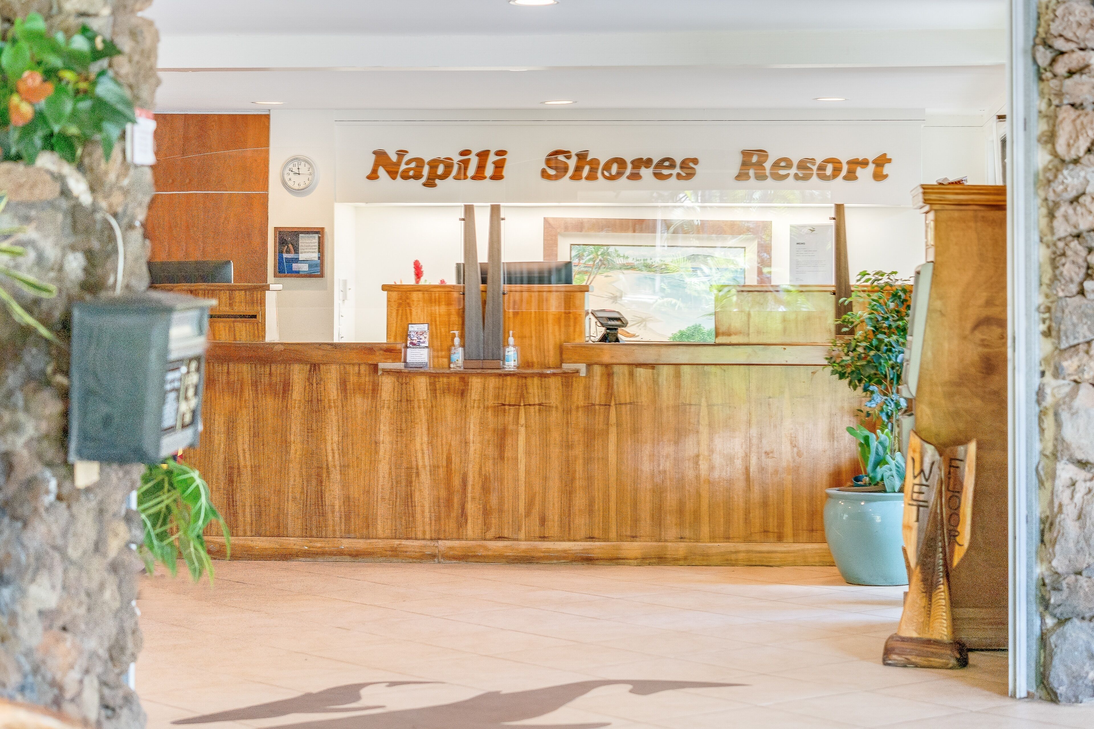 Un comptoir d’accueil en bois au Napili Shores Resort, avec une horloge et une vue sur l’océan par la fenêtre.