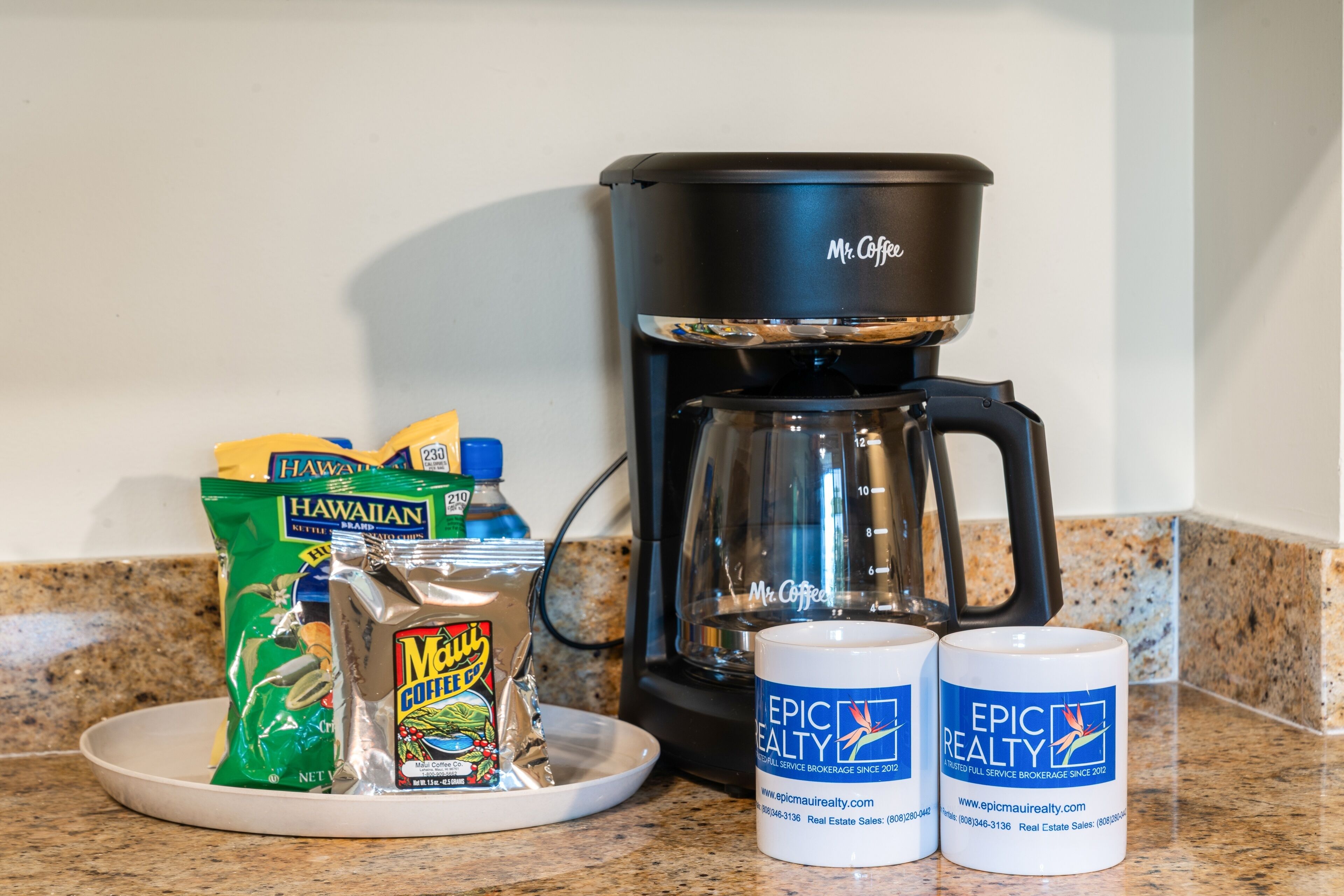 A kitchen counter with a Mr. Coffee coffee maker, a packet of Hawaiian coffee, a Mr. Coffee mug, and a Mr. Coffee mug with the Epic Realty logo.