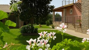 A building with a stone facade, a covered outdoor area with a metal railing, and a well-maintained lawn with flowers. - Comfy nest in a garden, 1,5km to the sandy beach (Marathopoli)