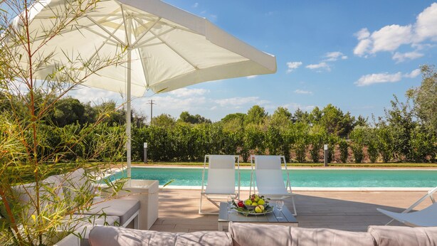 A poolside area with white lounge chairs, a table with fruit and a glass, and a large umbrella. - Villa Cavuzzi (Polignano a Mare)