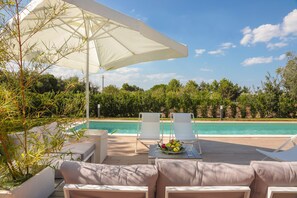 Zona de piscina con sillas reclinables blancas, una mesa con frutas y un vaso, y una sombrilla grande.