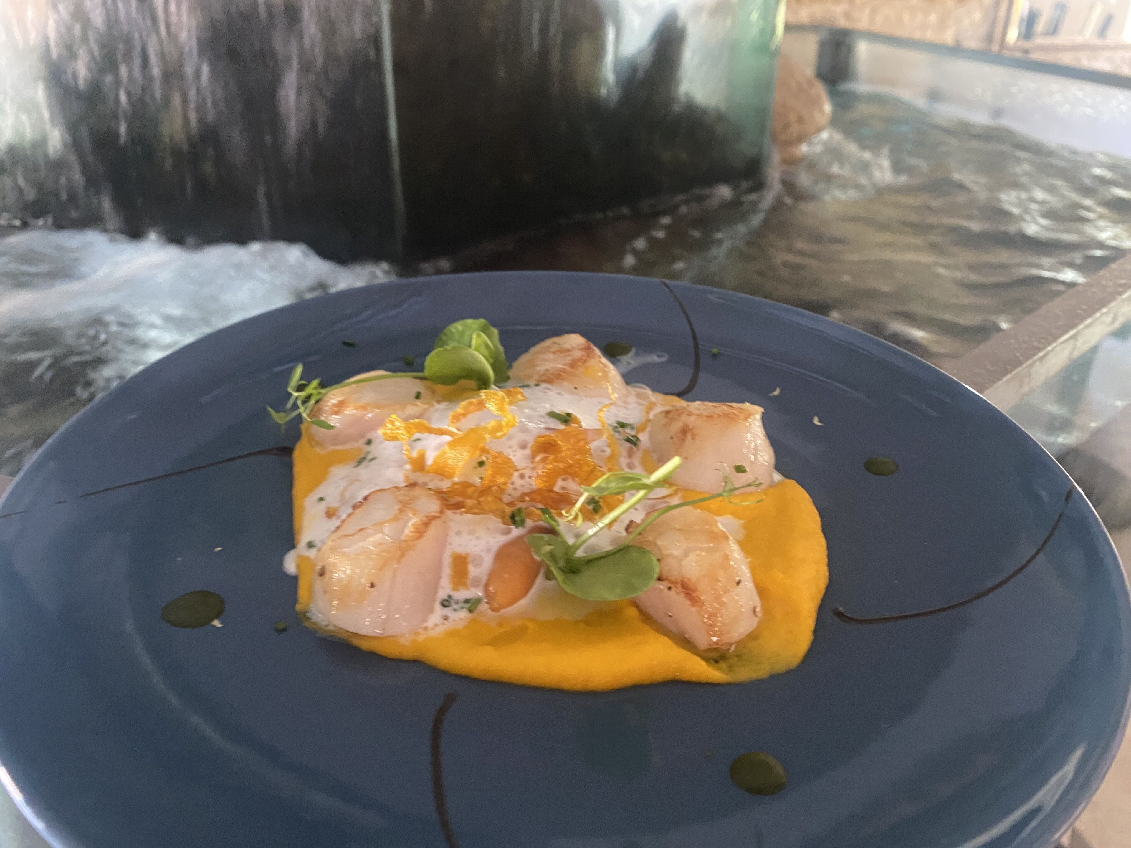A blue plate with a seafood dish, garnished with herbs and orange strips, on a glass table with a blurred background.