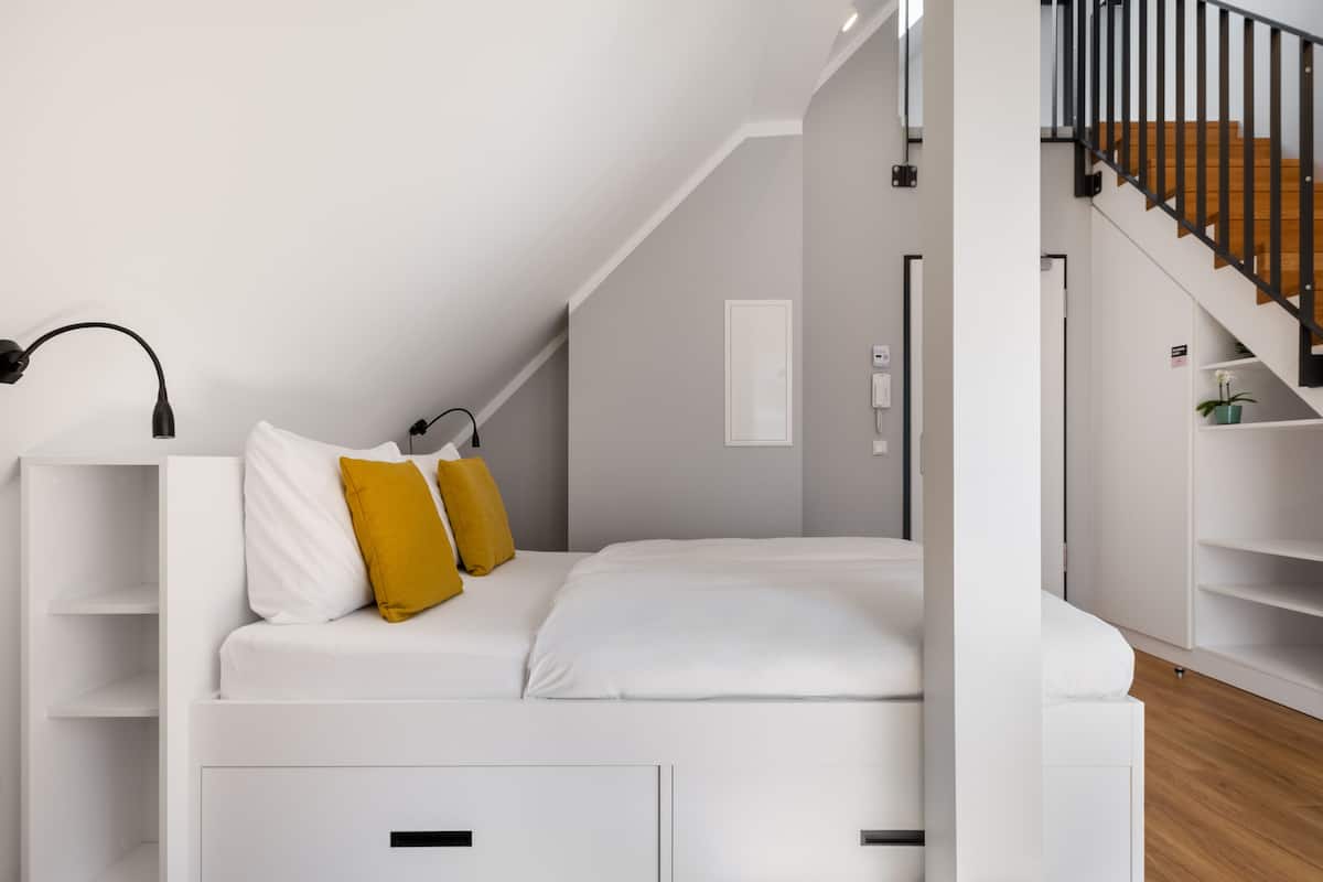 Une chambre de style loft moderne, avec un lit blanc, un plancher en bois et un escalier doté d’une rampe noire.
