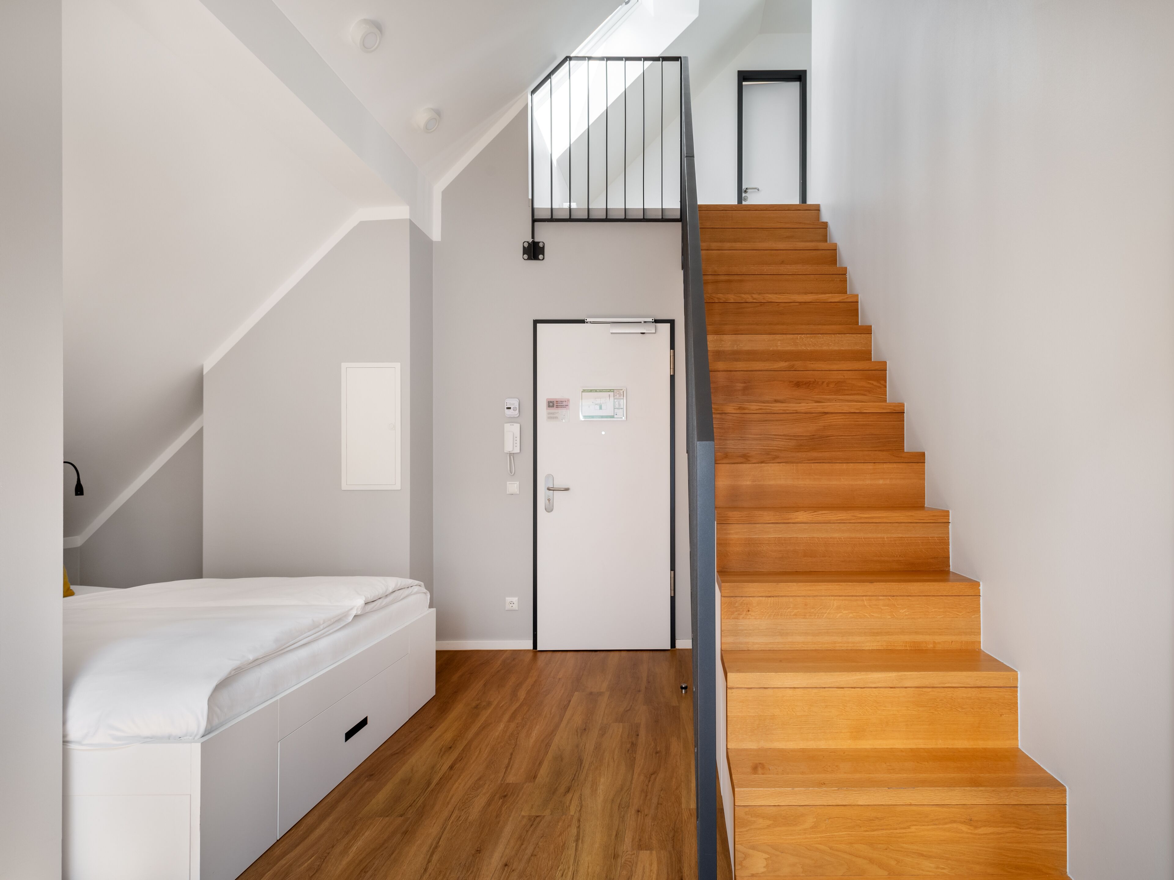 Een moderne slaapkamer met een houten trap, een bed en een deur met een lichtschakelaar en een thermostaat.